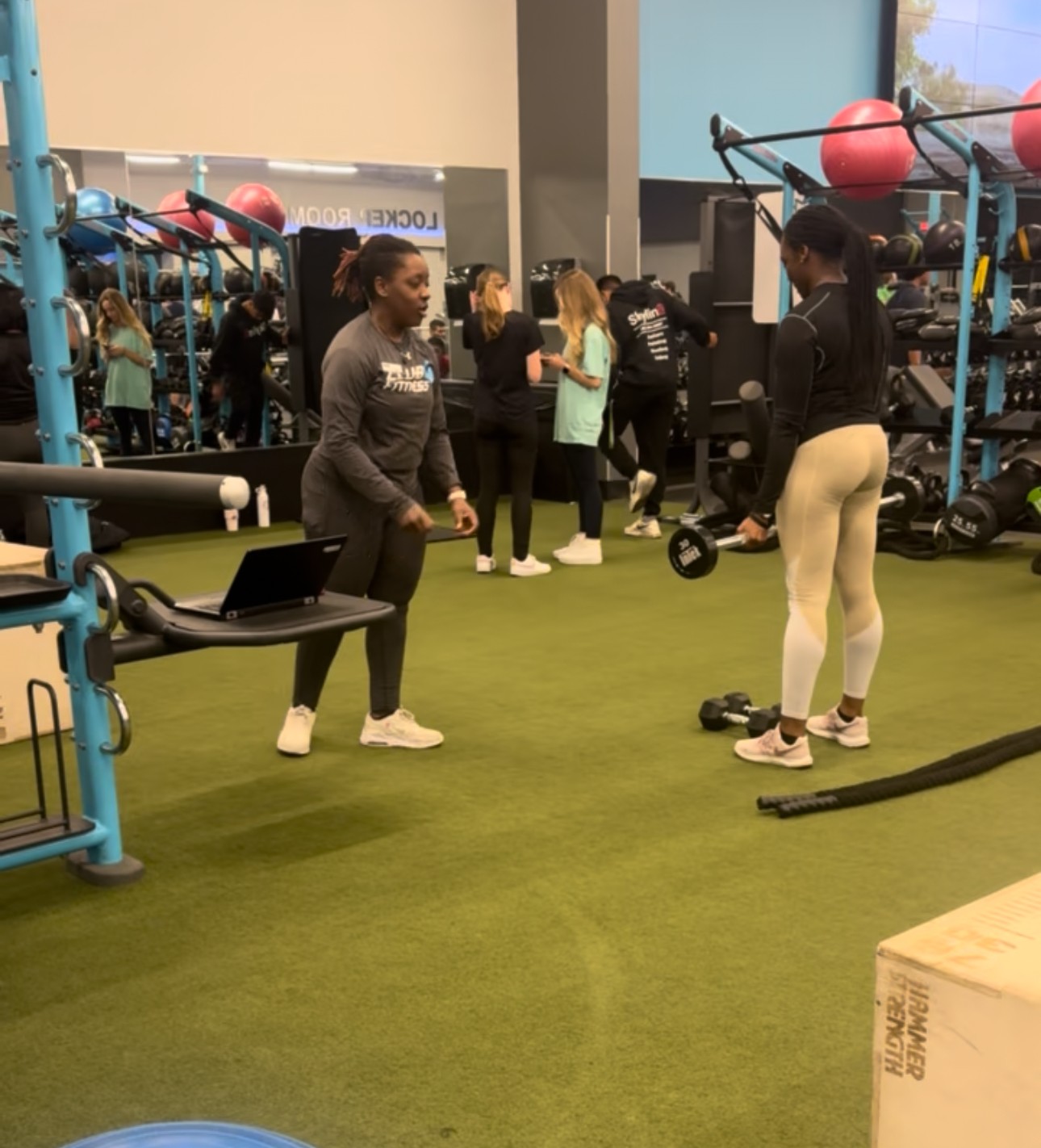Two women in workout clothes talking in a gym with exercise equipment and other people exercising in the background.