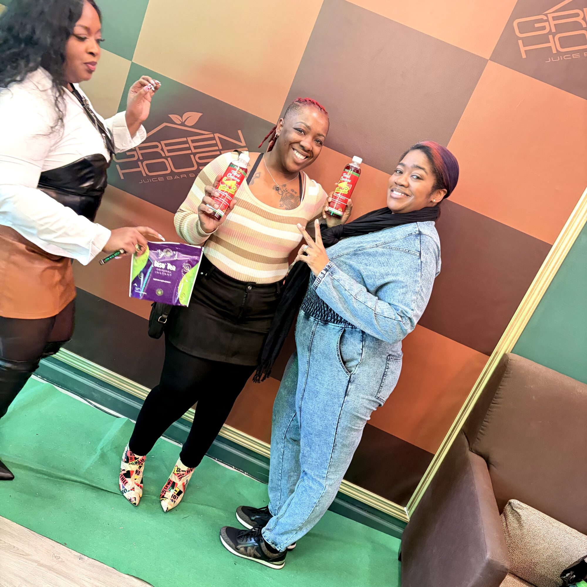 Three women smiling and holding products in front of a colorful backdrop, one woman pointing at the products.