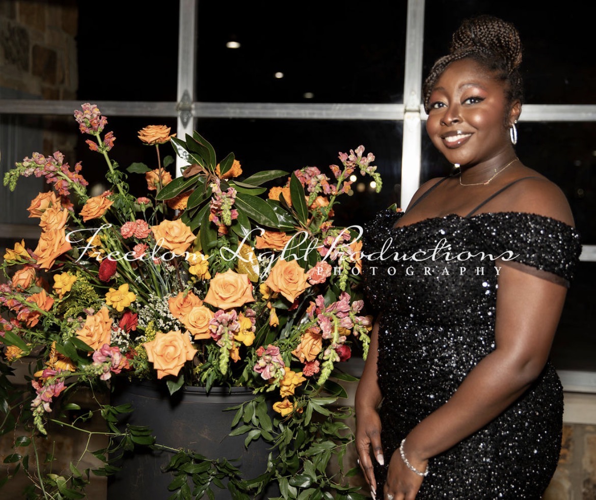 Woman in a black, sparkly off-shoulder dress standing next to a large colorful flower arrangement, smiling.