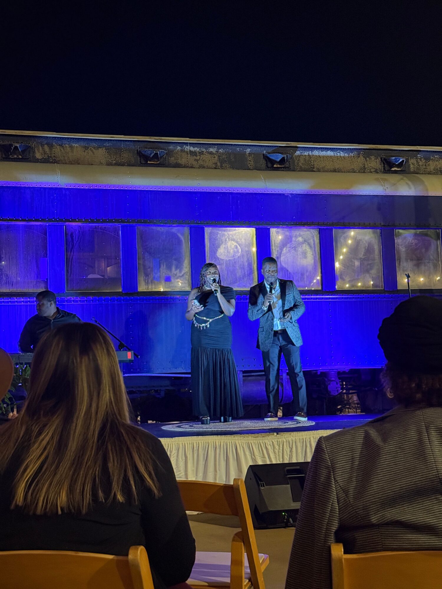 People on stage with blue lighting, some holding microphones, audience seated in front, woman with long hair in foreground.