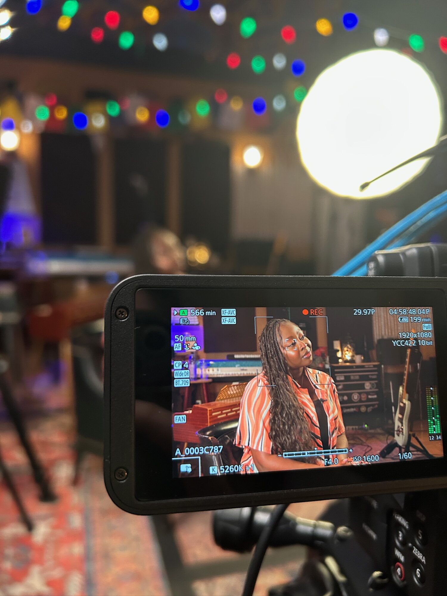 Person with long hair and striped shirt is being filmed in a studio with colorful lights and equipment in background.