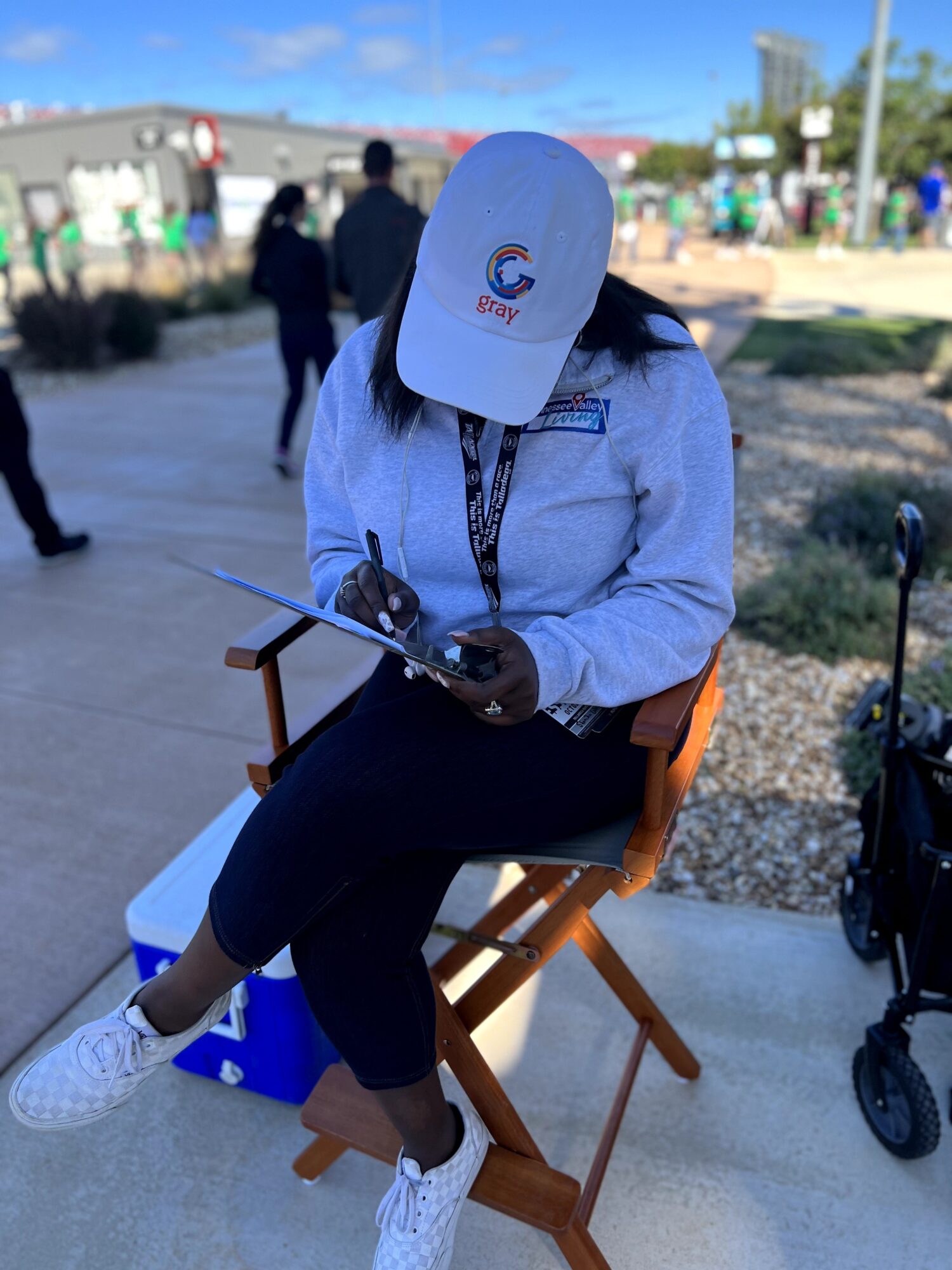 Person sitting on a wooden chair outdoors, wearing a white cap, light blue jacket, and black pants, looking at a phone.