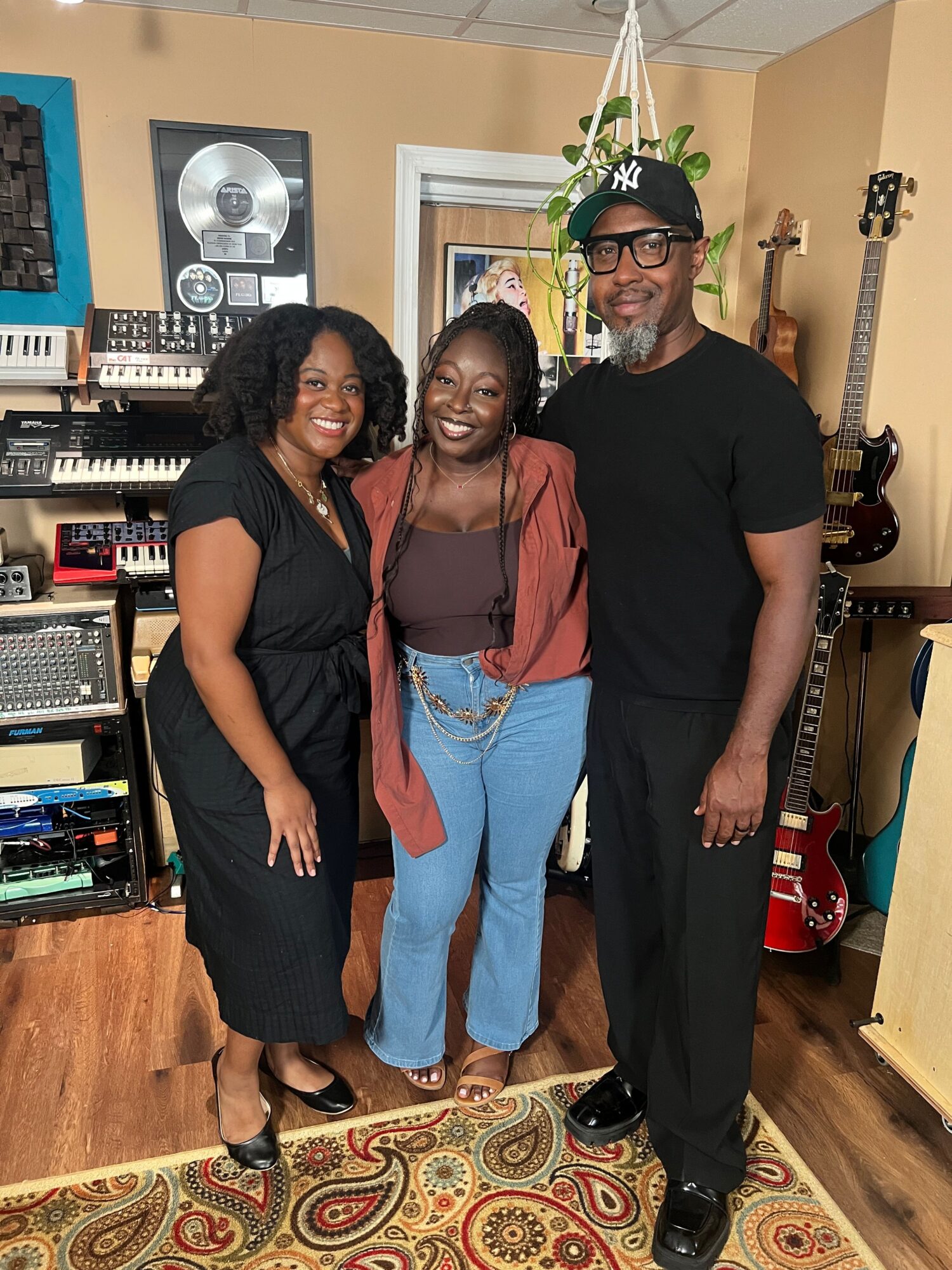 Three people standing in a room with musical instruments and equipment, smiling at the camera.