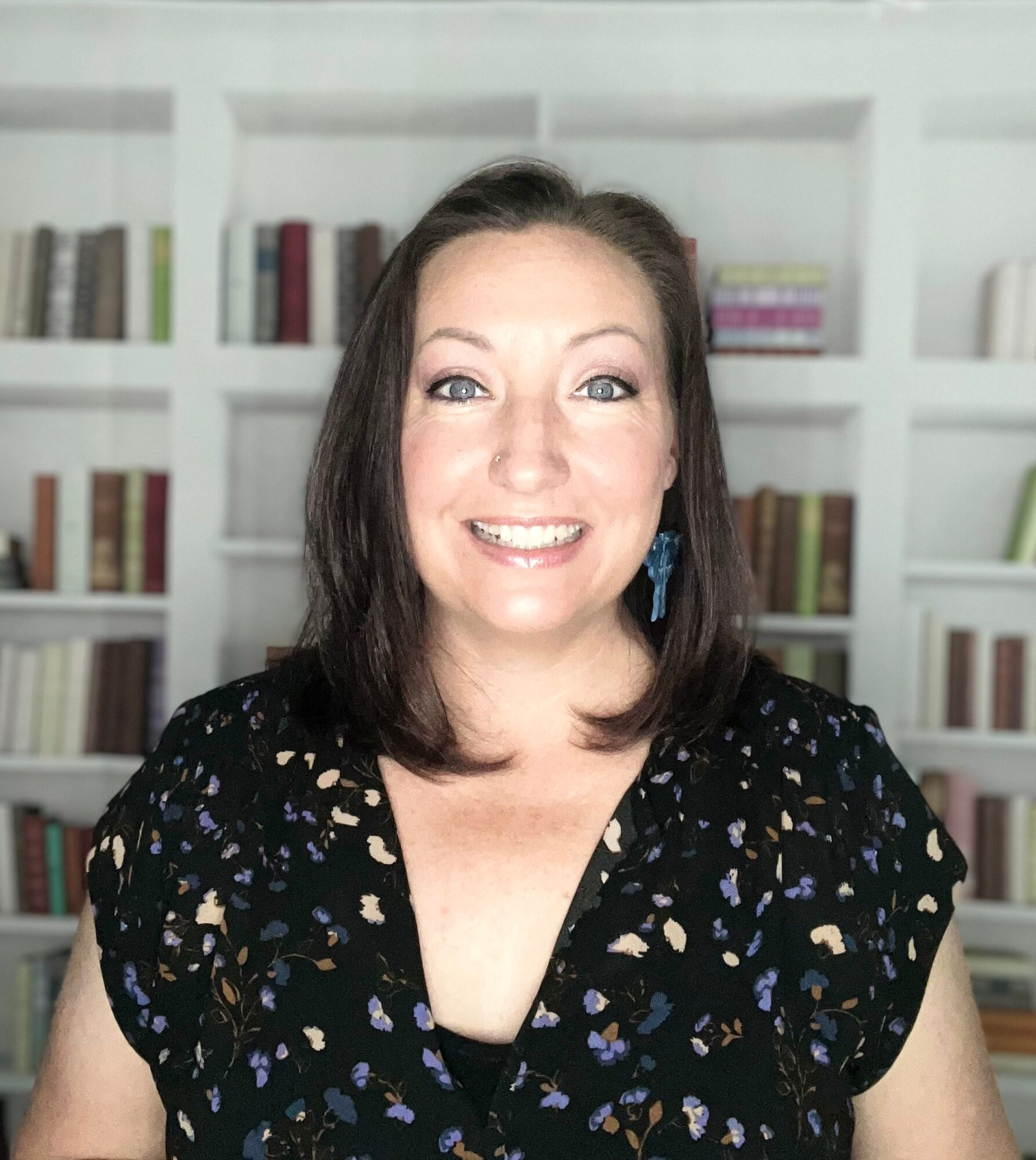 Smiling woman with shoulder-length dark hair, wearing a black patterned top, in front of a bookshelf background.