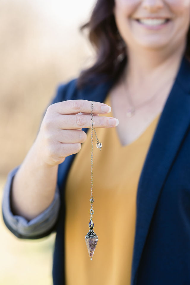 Person holding a necklace with a pendant, smiling, outdoors, wearing a yellow top and blue blazer.