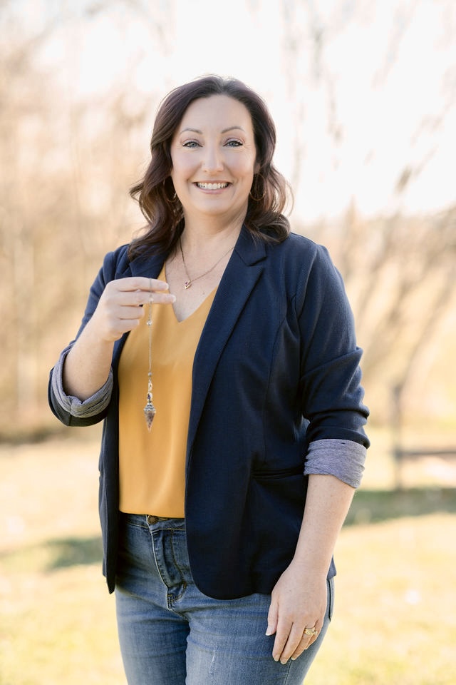 Smiling woman outdoors wearing a navy blazer and yellow top, holding a necklace, with blurred trees in background.