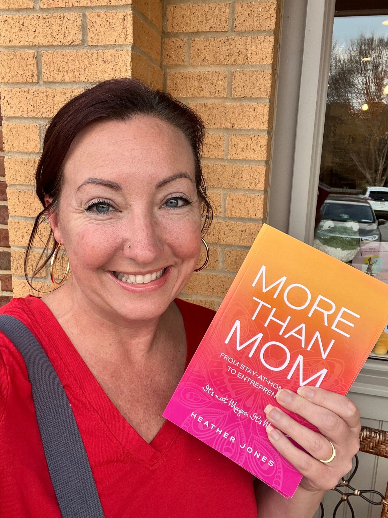Woman smiling holding a colorful book titled 'More Than Mom' outside near a window.