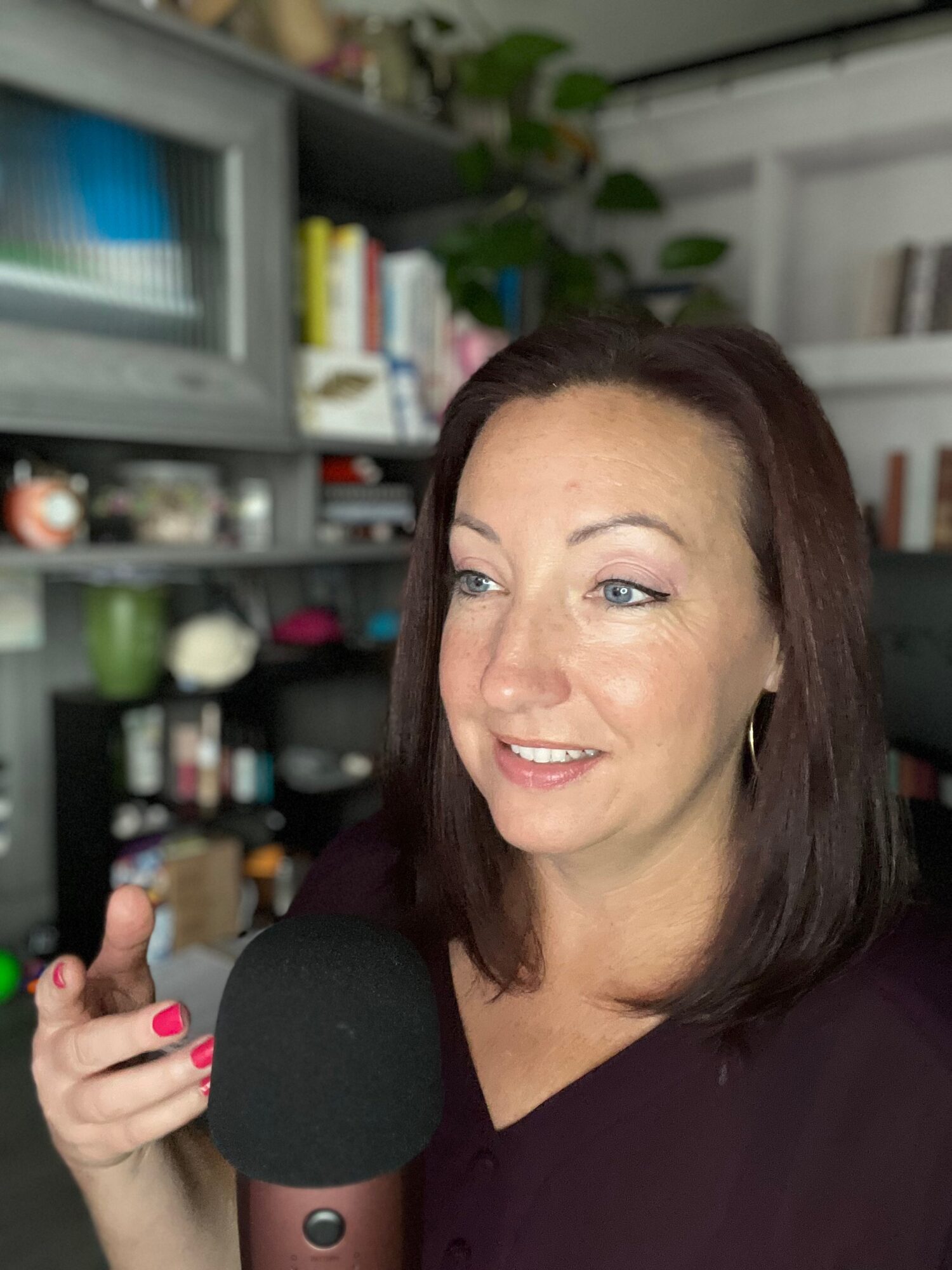 Woman with dark hair and earrings speaking into a microphone in a room with bookshelves.