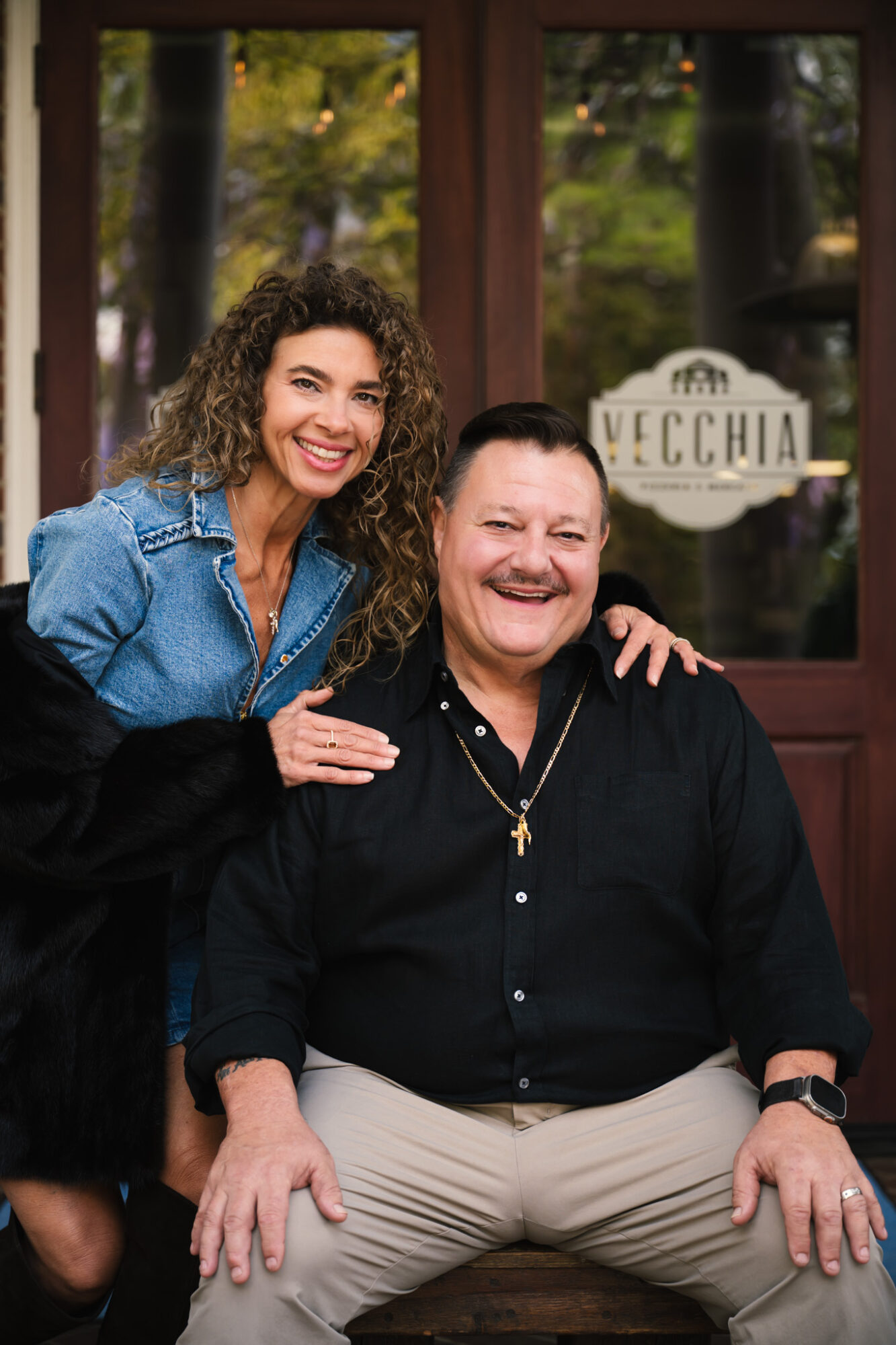 Two women smiling, one standing with arm around the other sitting outside a building with a sign that reads 'VECCHIA'.