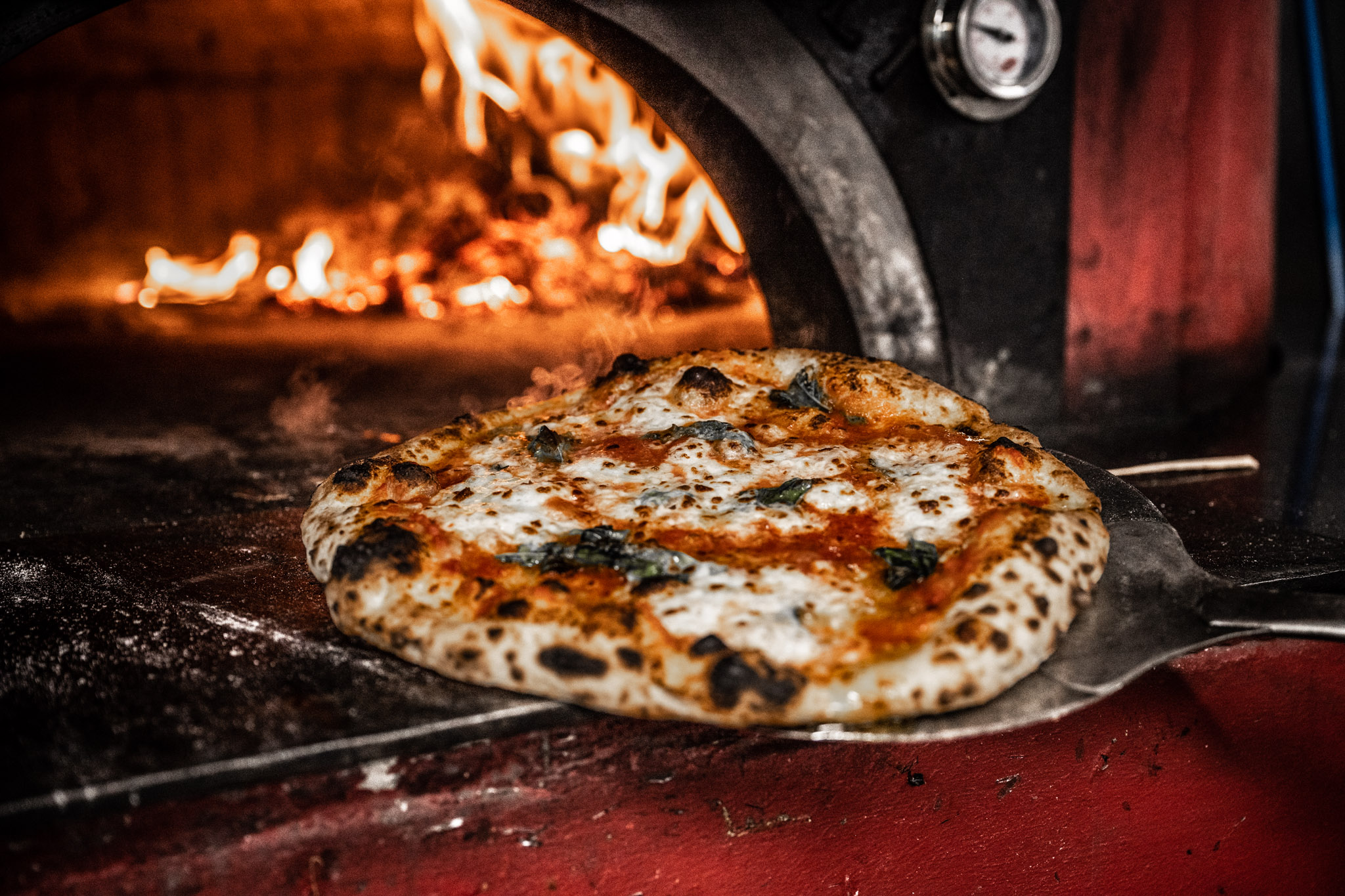 Pizza being cooked in a wood-fired oven with flames visible in the background.