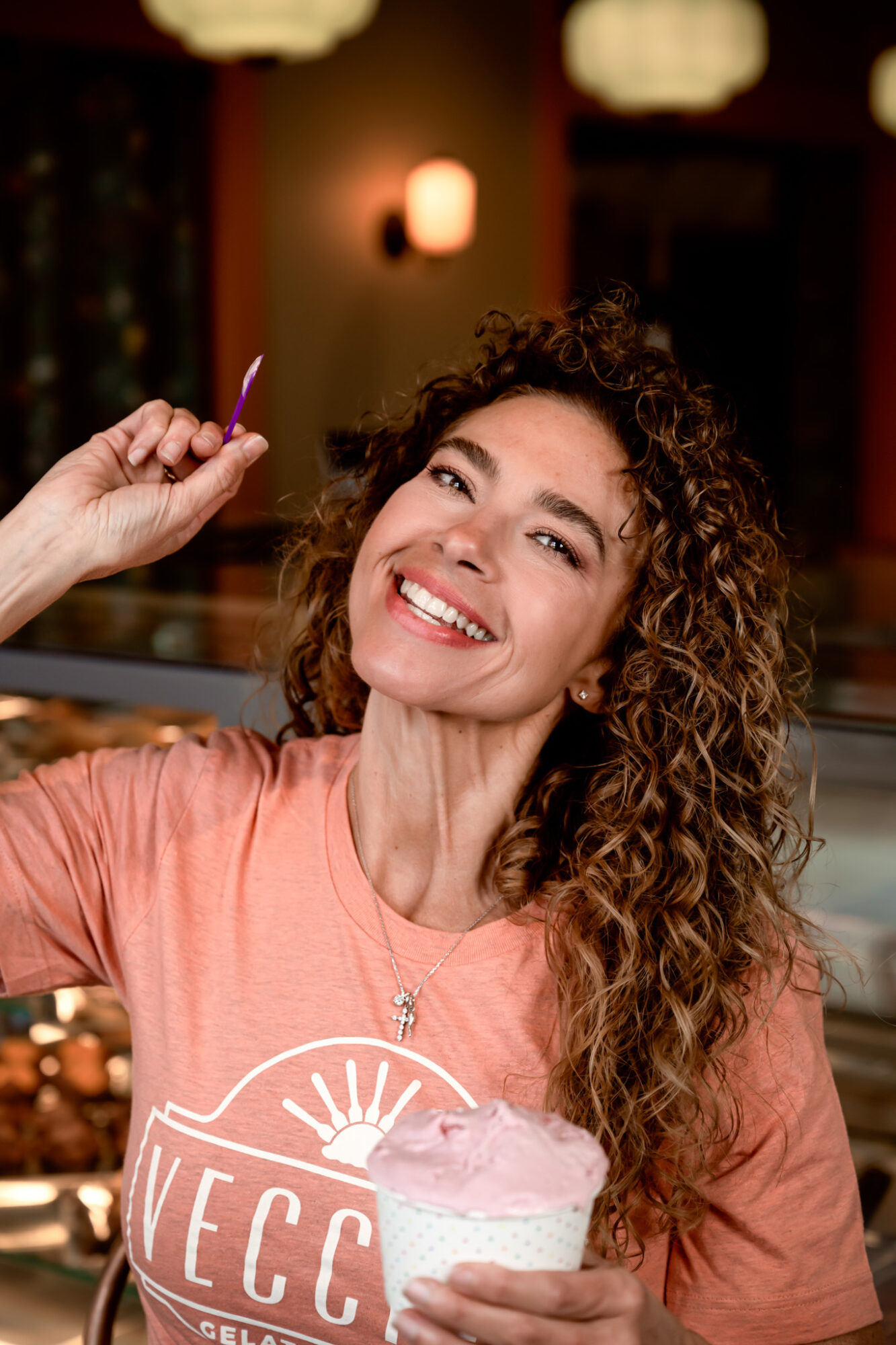Smiling woman with curly hair holding a pink-colored drink in a glass, indoors with warm lighting.