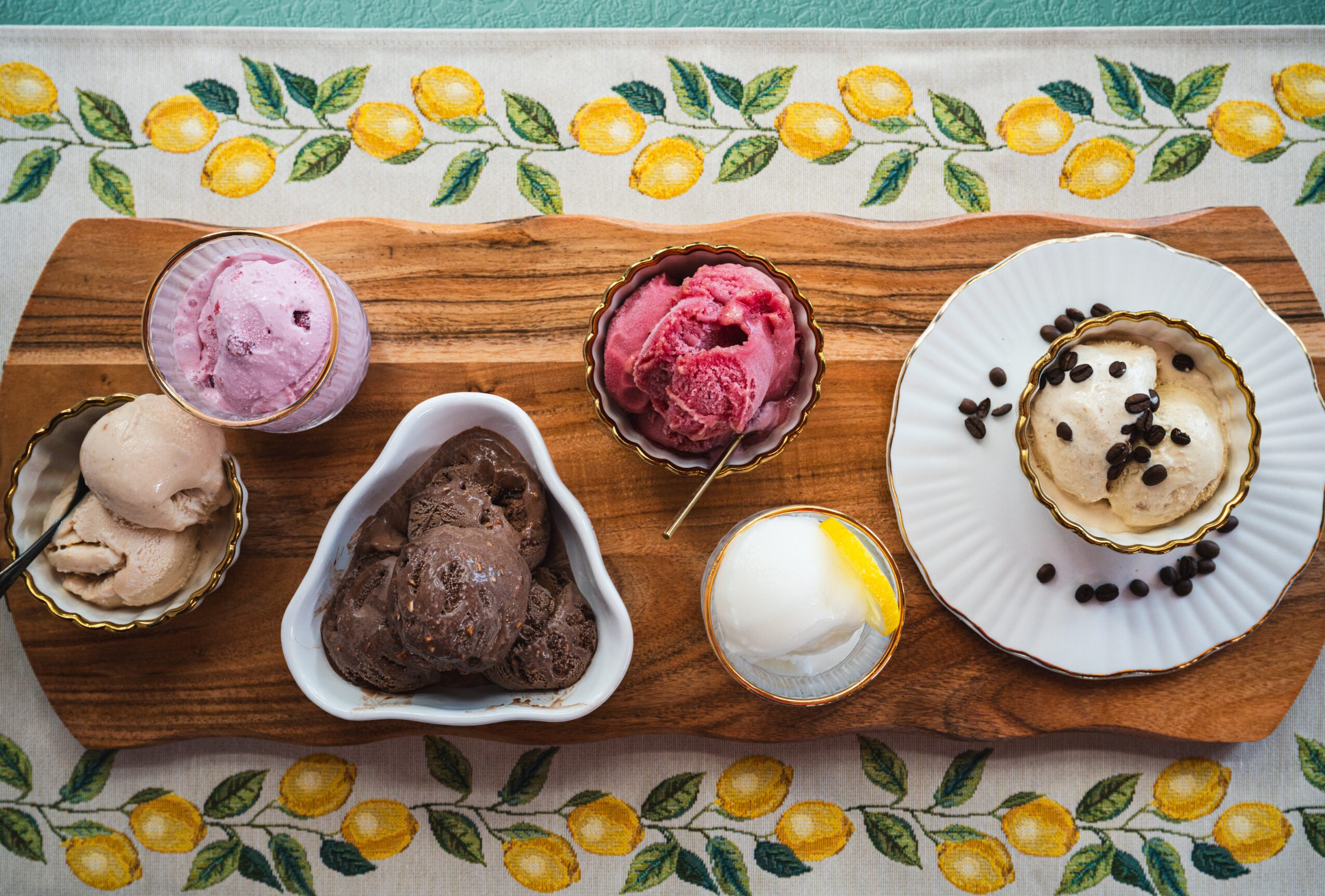 Assorted desserts on a wooden board with lemon-patterned tablecloth, including ice cream, cake, and mousse, with a spoon and berries.