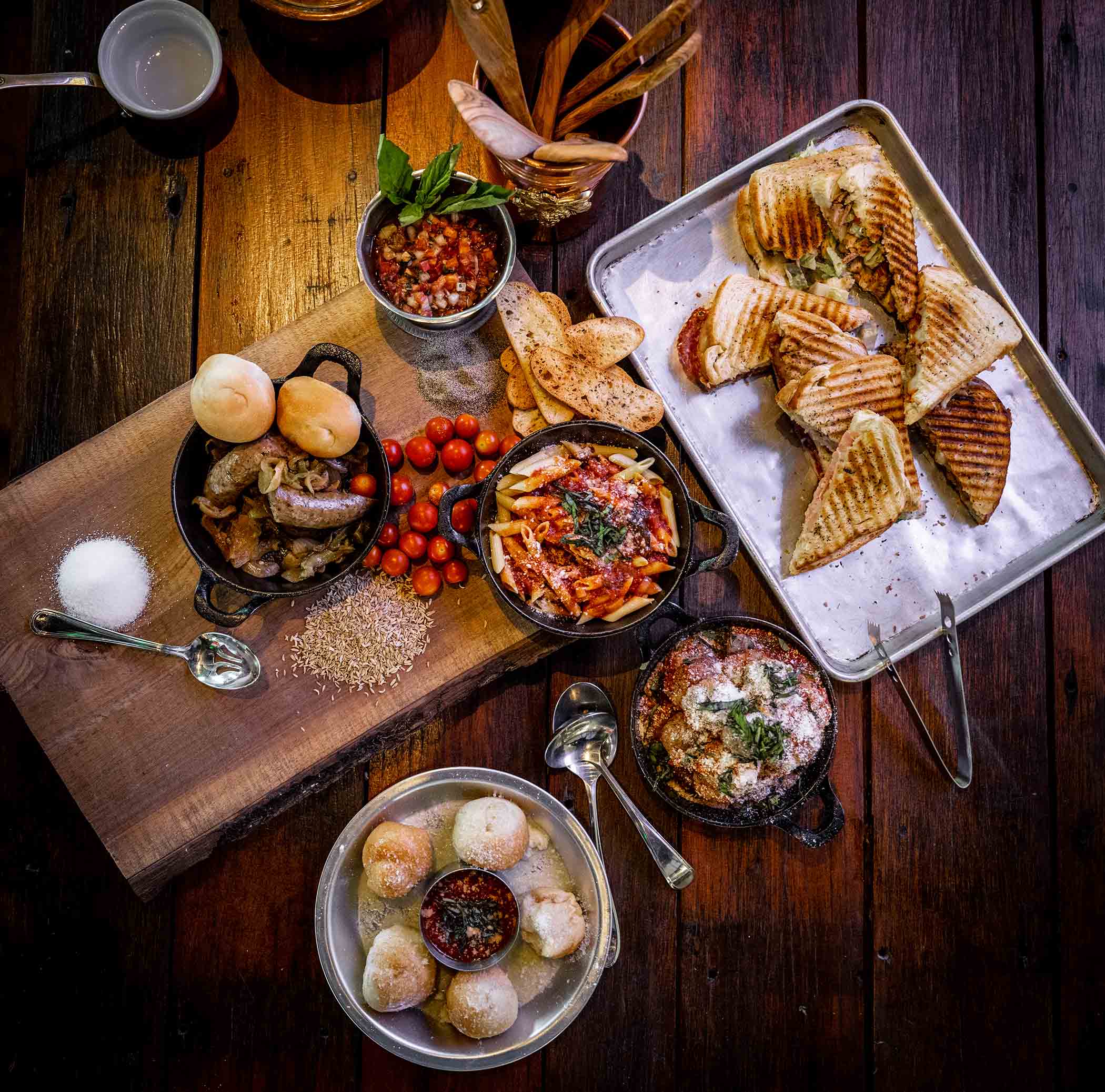 Assorted food including grilled sandwiches, cherry tomatoes, bread, and a bowl of dumplings on a wooden table.