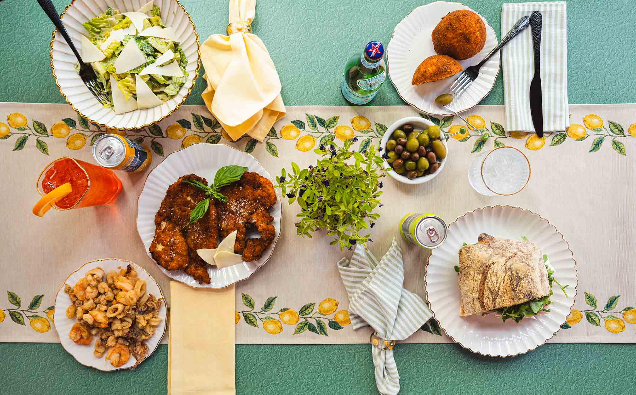Table with various dishes, including fried chicken, salad, bread, and drinks, on a tablecloth with a lemon pattern.