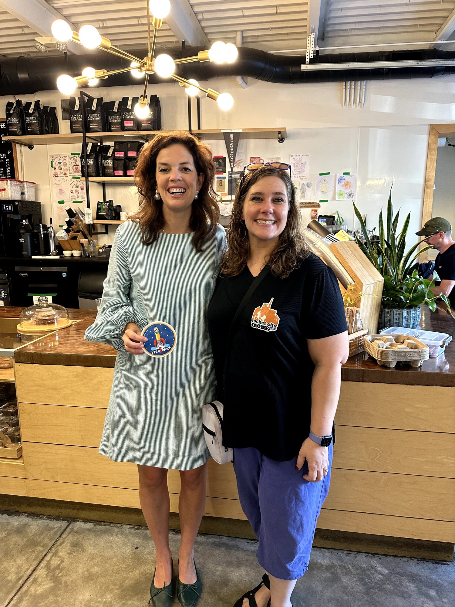 Two women stand inside a coffee shop, smiling, with a counter and shelves behind them.