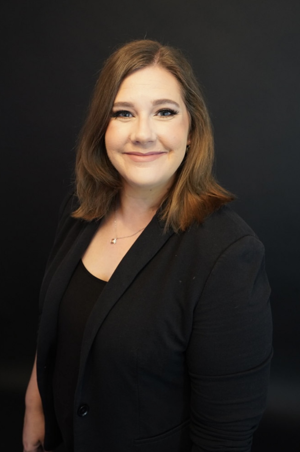 Portrait of a woman with shoulder-length brown hair, smiling, wearing a black blazer and top, against a dark background.