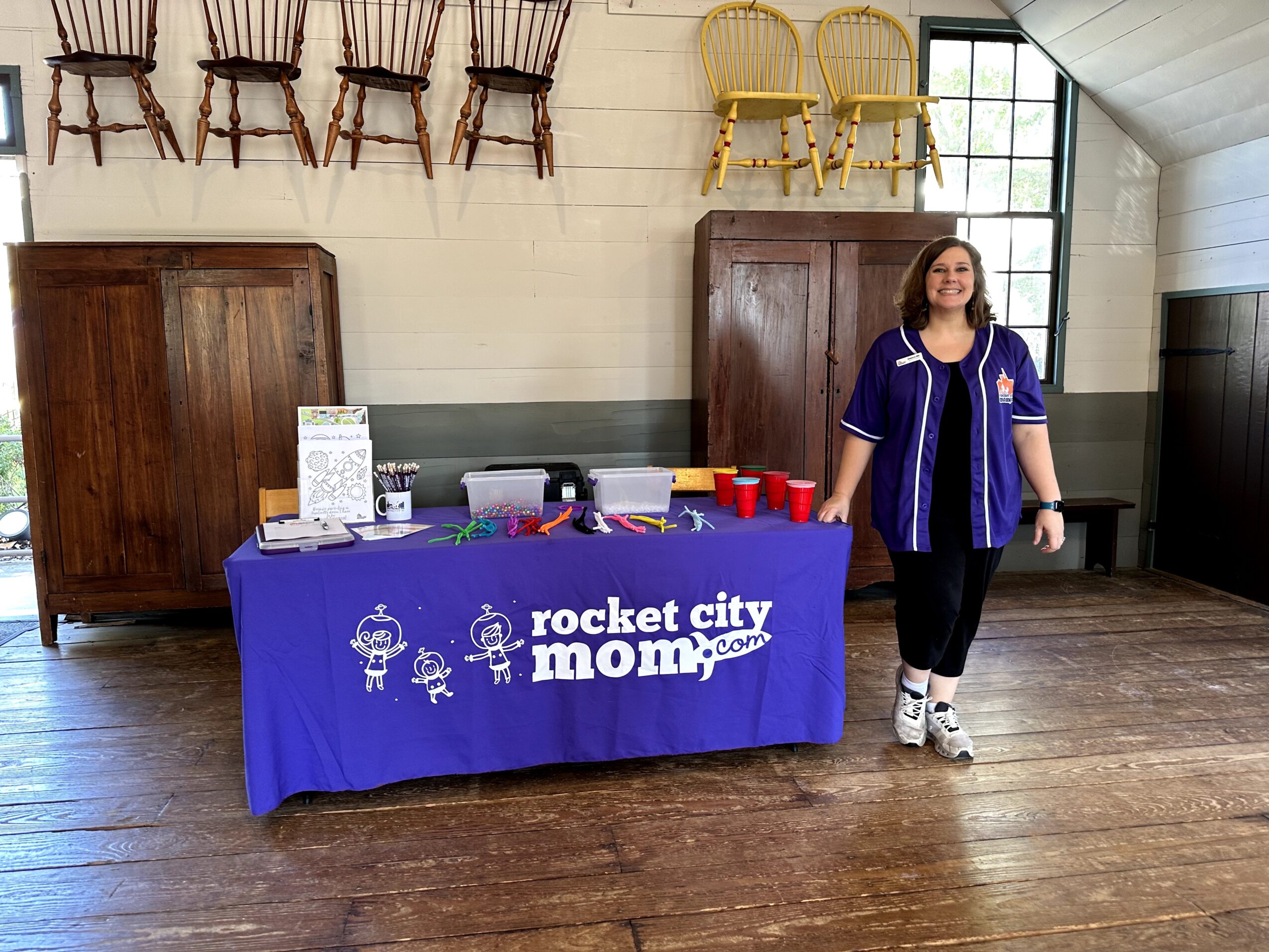 Woman standing next to a table with a blue tablecloth that reads 'rocket city mom' in a room with wooden floors and chairs on the wall.