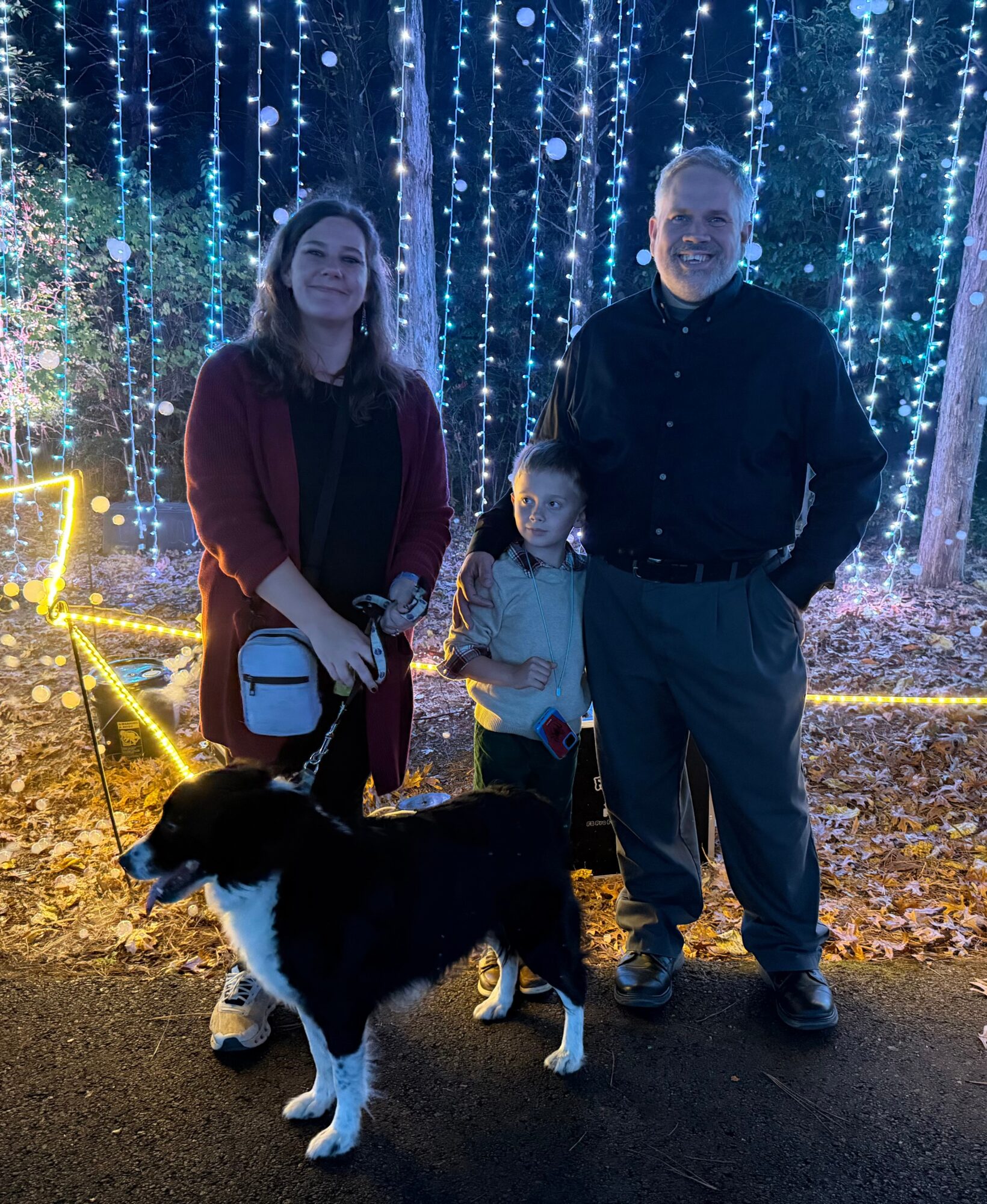 Three people and a dog outdoors at night with blue string lights and fallen leaves, smiling at the camera.