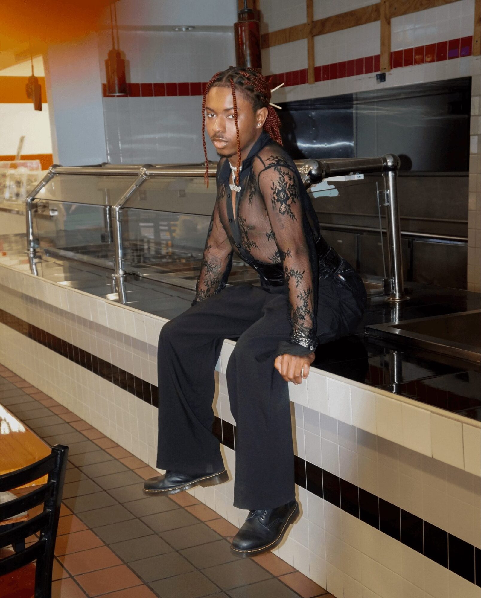 Person with braided hair in a black lace top and wide-leg pants sitting on a counter in a restaurant or cafe, with a glass display case behind them.