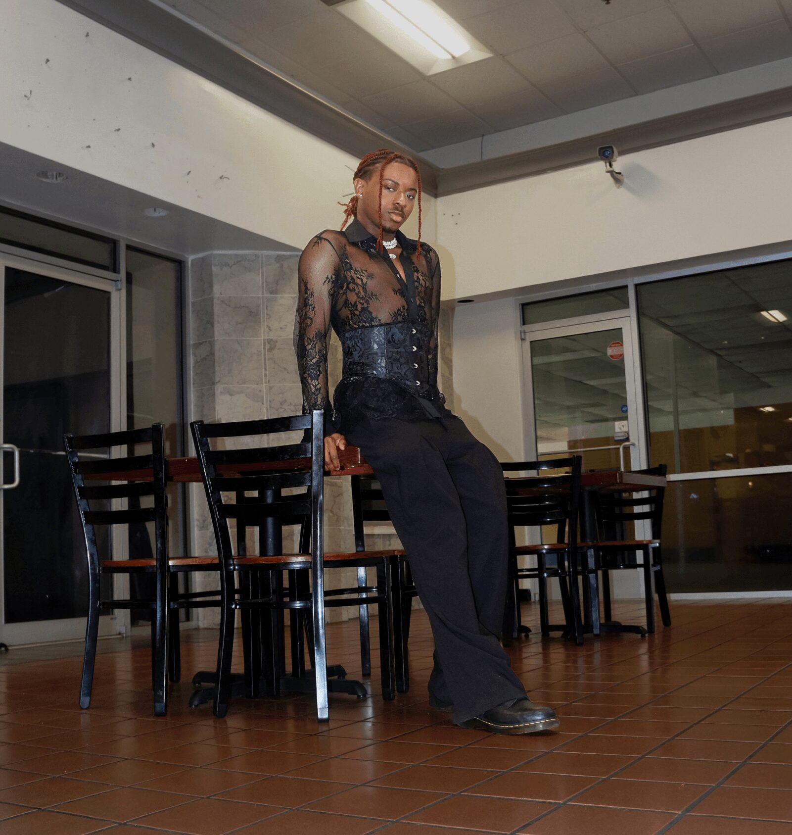 Person sitting on a table in a room with chairs, wearing a sheer black top and black pants.