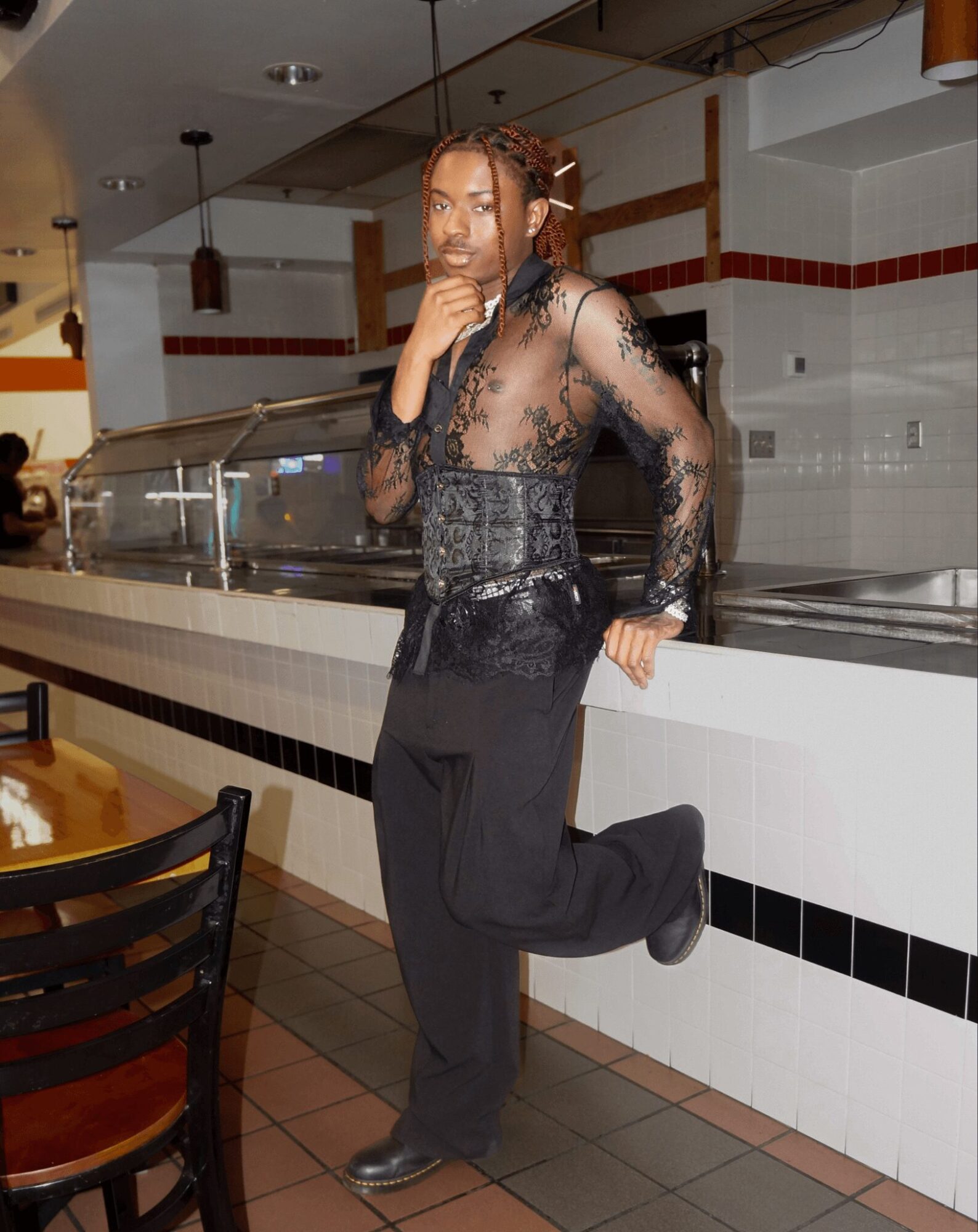 Woman in black lace top and pants standing in a restaurant, one leg raised, hand near chin, looking at camera.