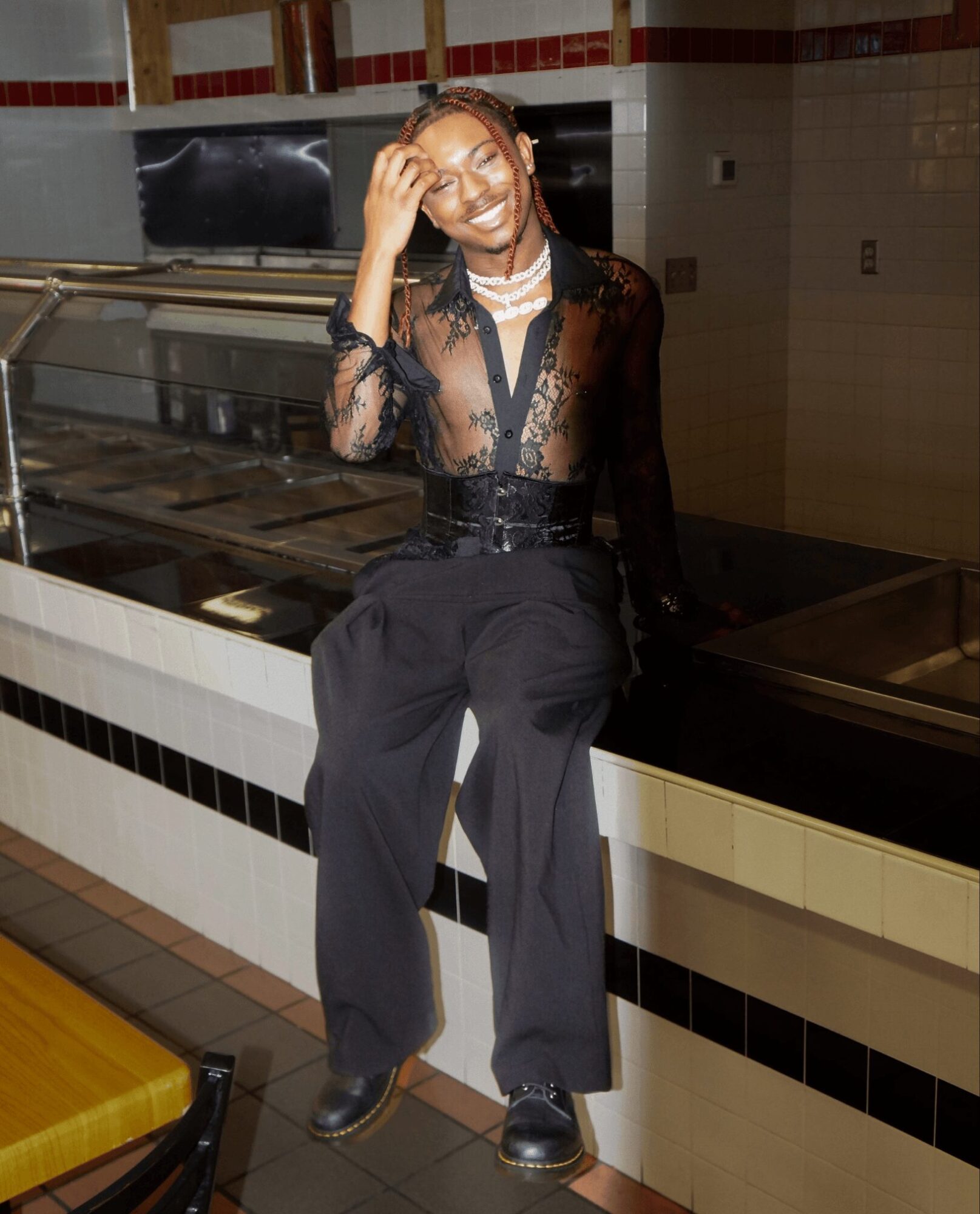 Person sitting on a counter, smiling, wearing a sheer black top and black pants, in a tiled room with a yellow seat nearby.