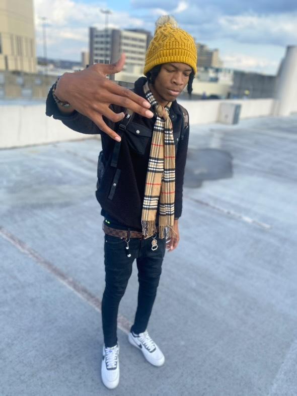 Young person wearing a yellow knit hat, black jacket, plaid scarf, jeans, and white sneakers on rooftop with cityscape background.