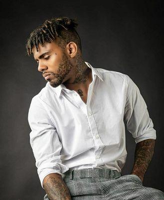 Man with dreadlocks and tattoos wearing a white shirt and gray pants, looking down, against a dark background.