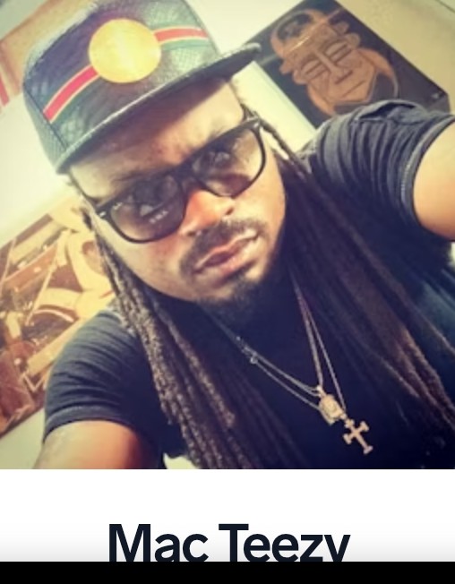 Person wearing glasses, a colorful hat, and multiple necklaces, taking a selfie indoors with artwork in the background.