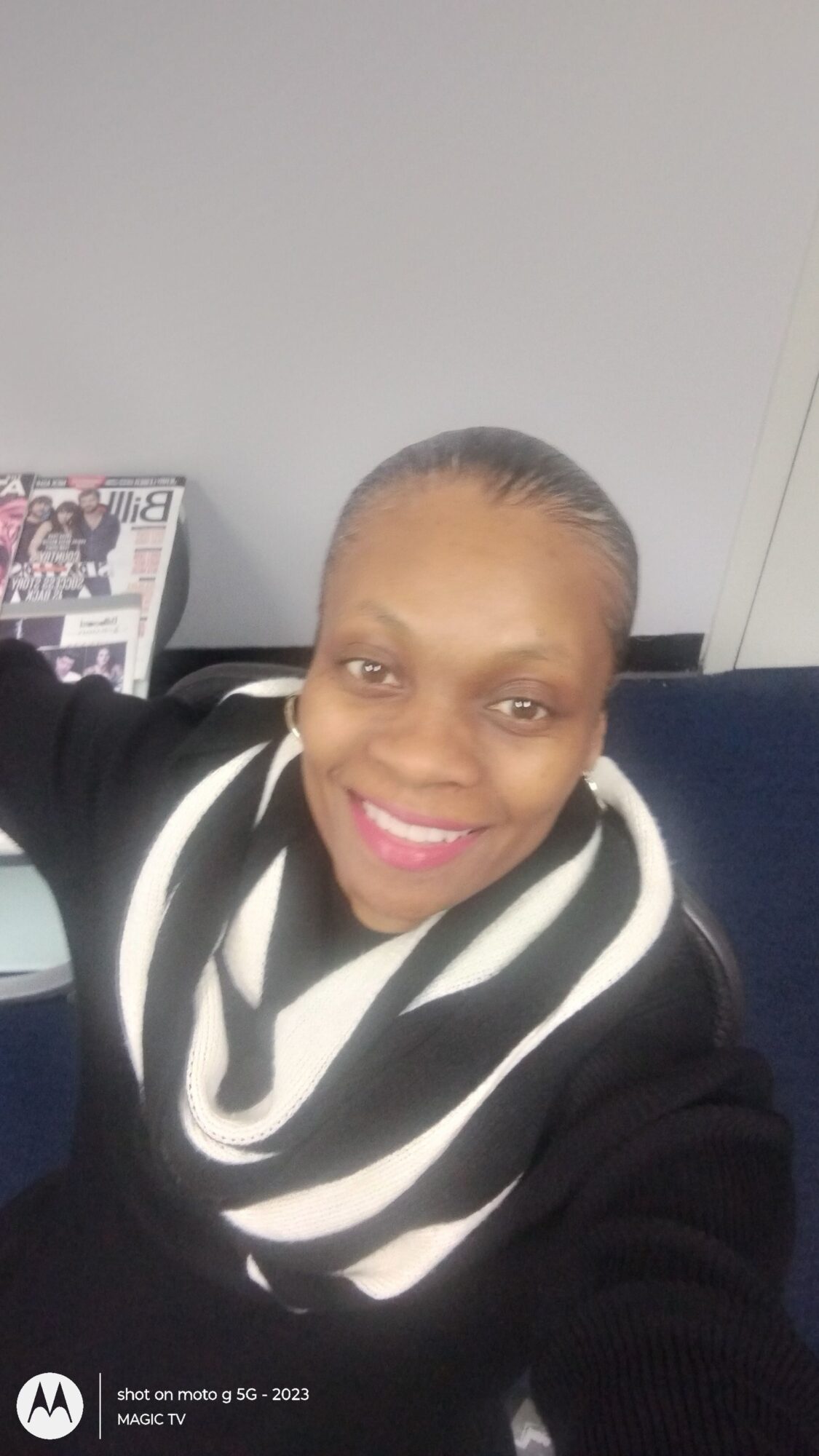 Woman with braided hair smiling, wearing a black and white striped scarf, sitting indoors against a gray wall.