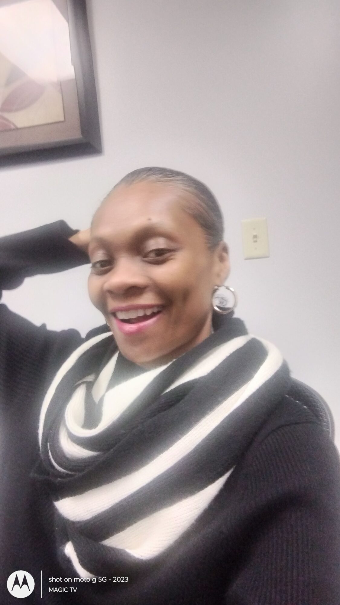 Woman with a bun hairstyle smiling, wearing a black and white striped scarf and black top, sitting indoors.