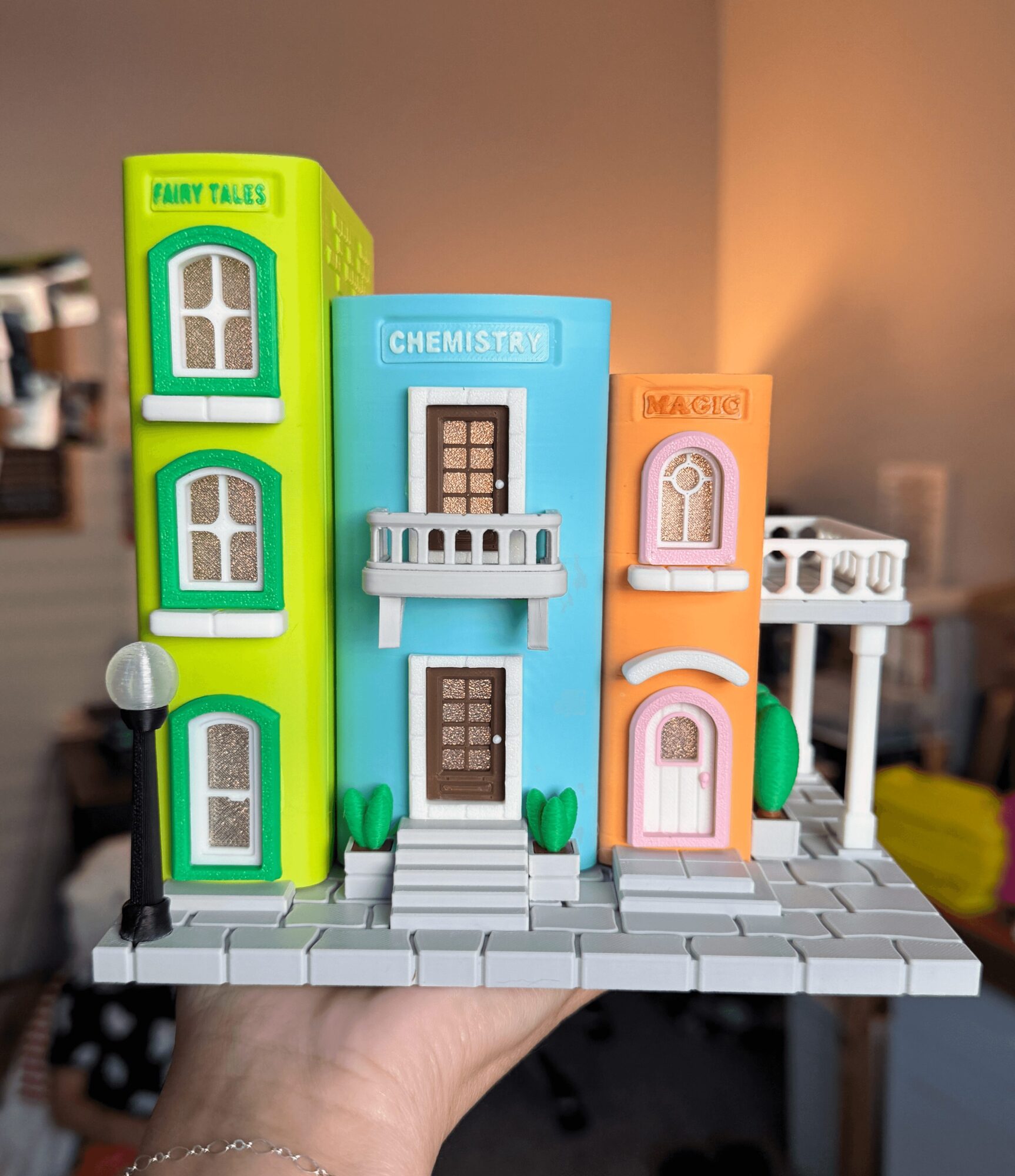 Hand holding a miniature cityscape with colorful toy buildings labeled 'Fairy Tales,' 'Chemist,' and 'Magic,' on a gray base.