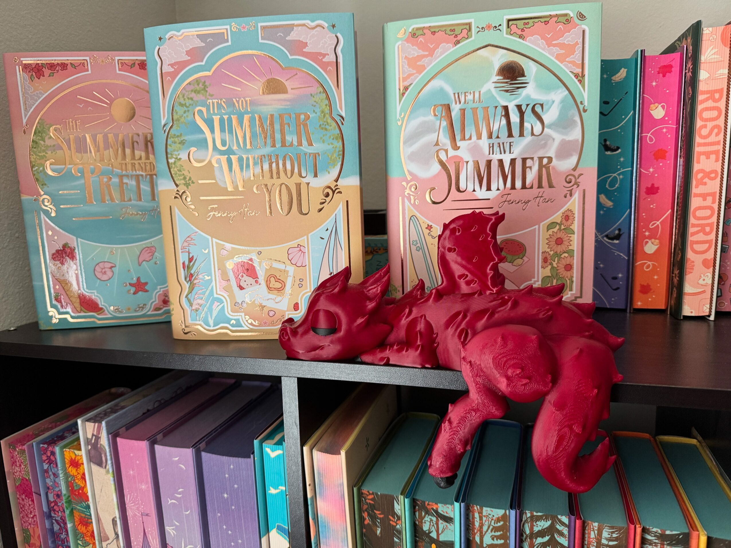 Pink plush unicorn toy lying on a shelf with colorful summer-themed books behind it.
