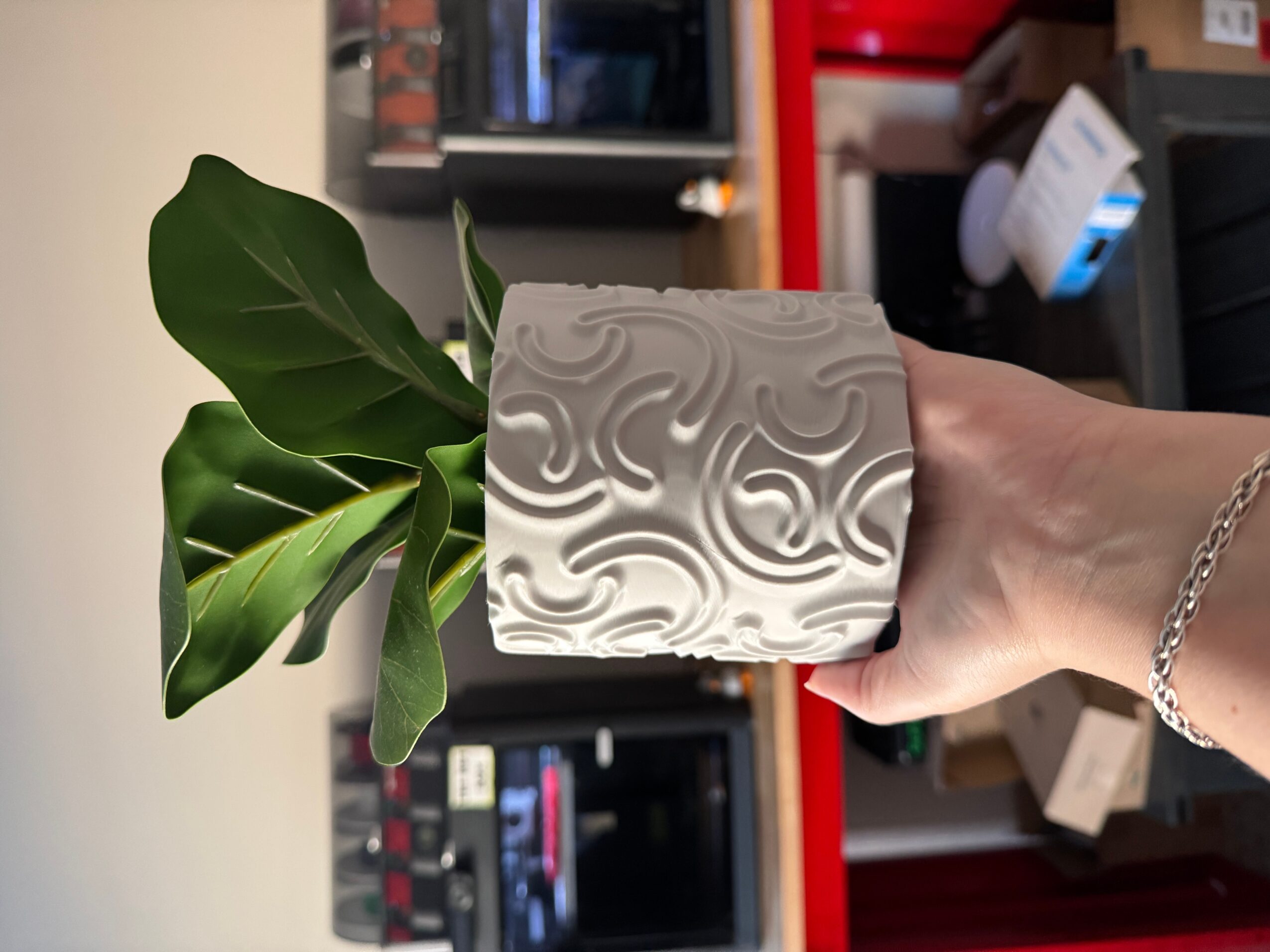 Hand holding a decorative white cube with swirling patterns, with a green plant in the background, in an indoor setting.