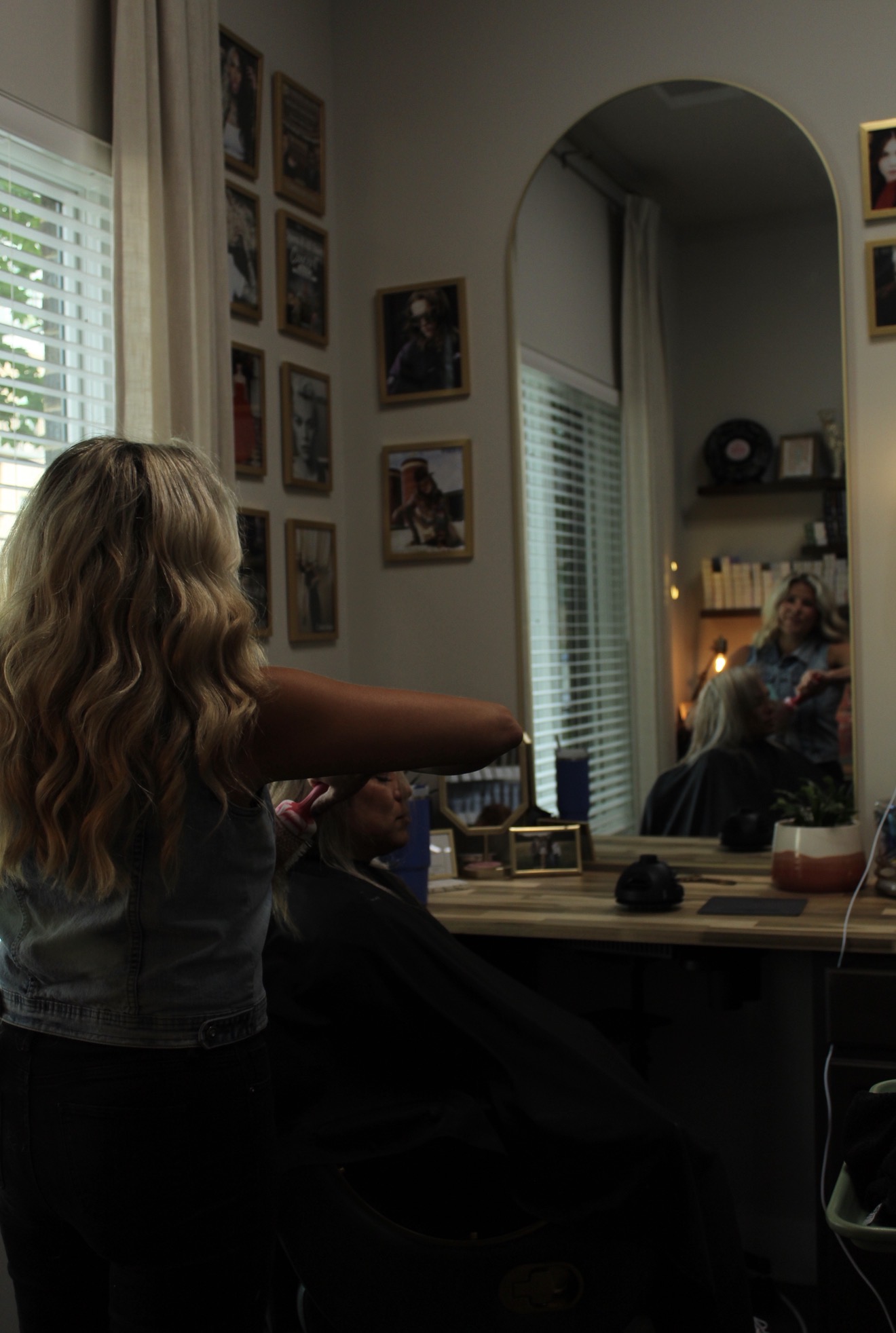 Child with long hair reaching towards a mirror in a room with framed pictures, a window with blinds, and a person sitting in the reflection.