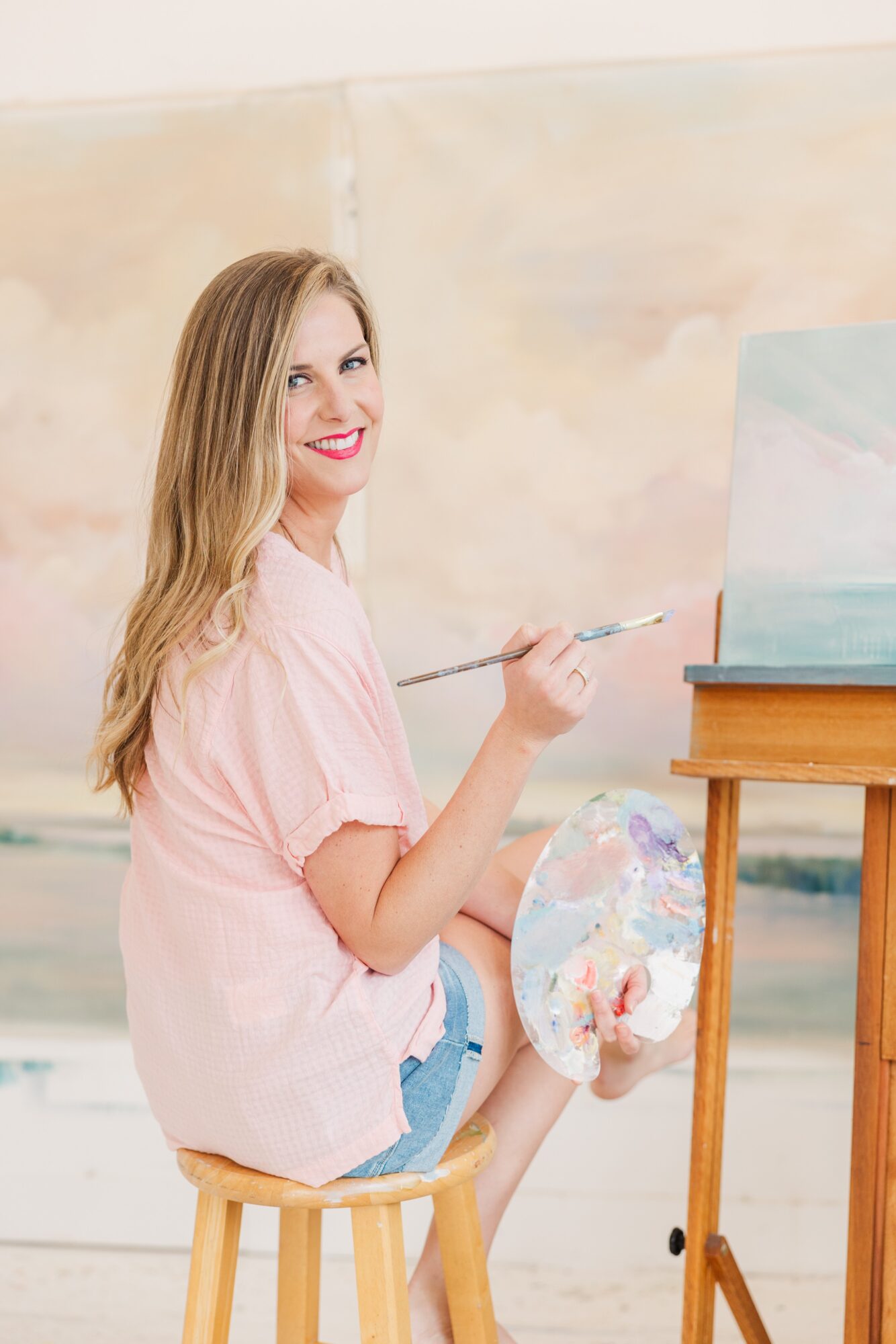 Woman with long hair sitting on a stool, holding a paintbrush and palette, smiling at the camera, painting on canvas.