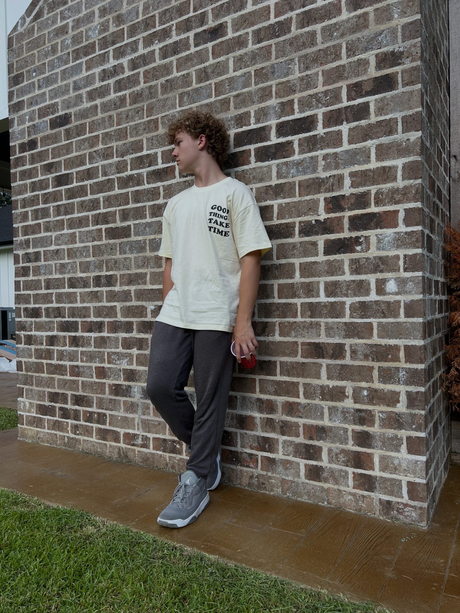 Person leaning against a brick wall outdoors, wearing a white T-shirt and dark pants, holding a red object.